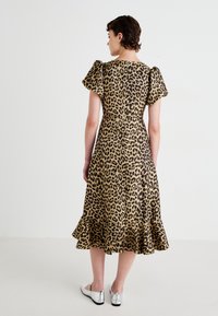 Žena v midi šatách s leopardím vzorom, s naberacími rukávmi a volánovým lemom, v kombinácii so striebornými plochými topánkami, stojí pred jednoduchým pozadím.