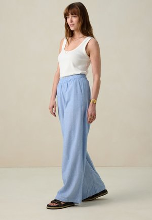 Pantalons larges texturés bleu clair avec une taille élastique. Portés avec un débardeur blanc et des sandales noires à design tissé.