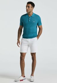 Teal gebreide polo met korte mouwen, patroontextuur en kraag; gecombineerd met witte shorts en casual sneakers met rode en blauwe accenten.