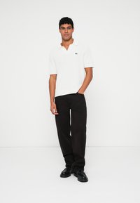 Polo shirt blanc texturé avec un col, des manches courtes, et un logo vert sur la poitrine, associé à un pantalon noir et des chaussures noires.