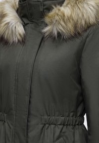 Manteau d'hiver vert foncé avec taille élastique et bordure en fourrure beige autour du col.