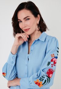 Light blue button-up shirt with puffed sleeves, featuring colorful floral embroidery on the sleeves and a soft, smooth texture.