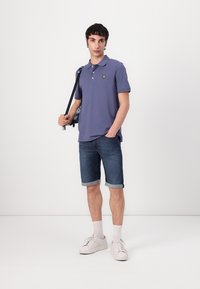 Violet polo marškiniai su geltonu logotipu, derinami su suvertais džinsiniais šortais ir baltais sportbačiais, stovintys prieš paprastą foną.