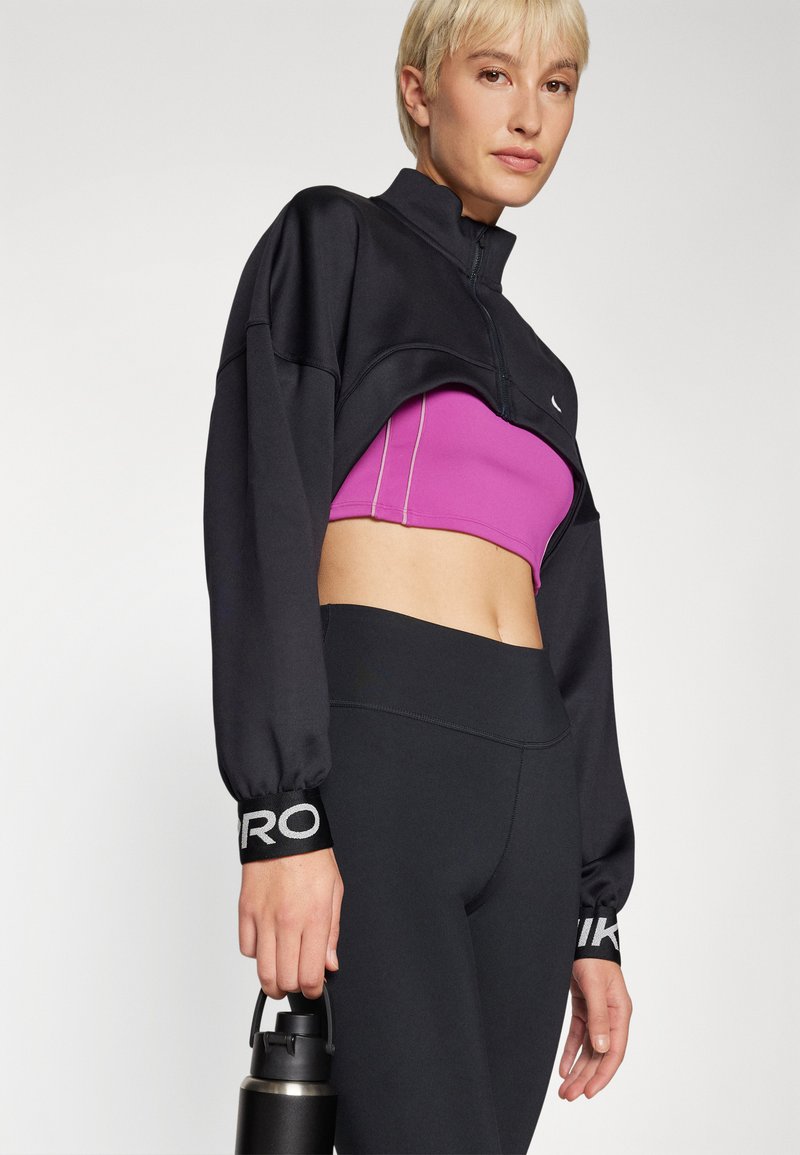 Schwarzer, kurzer Pullover mit hohem Kragen, kombiniert mit einem rosa Crop-Top und schwarzen Leggings. Enthält eine Wasserflasche in einer Hand.