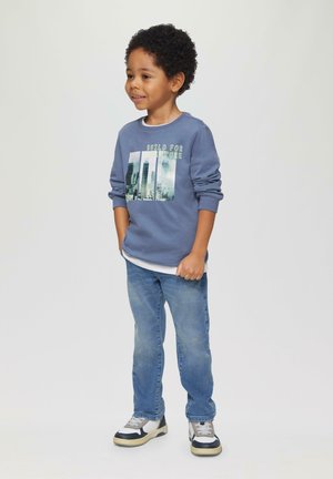 Blauer Sweatshirt mit dem Aufdruck "Built for Adventure", weißes Unterhemd, hellblaue Jeans und mehrfarbige Sneakers, stehend vor einem neutralen Hintergrund.