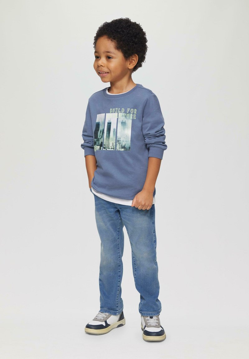 Blauwe sweatshirt met de tekst "Build for Adventure" grafisch ontwerp, witte onderhemd, lichtblauwe spijkerbroek en meerkleurige sneakers, staande op een neutrale achtergrond.