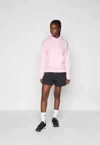 Hoodie rose avec une poche avant, associé à un short noir. Présente une coupe décontractée, des manches longues et une capuche avec cordon de serrage. Baskets noires et chaussettes blanches.