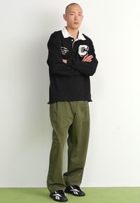 Polo nero a maniche lunghe con colletto bianco e logo "C", abbinato a pantaloni verdi oliva a vestibilità ampia e scarpe nere slip-on. Design semplice.