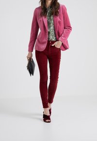 Růžový sametový blazer, květinová zelená košile, vínové kordové kalhoty a vínové sandály na podpatku, doplněné černou kabelkou.