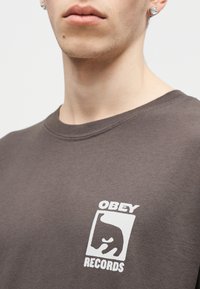 Κοντινό πλάνο ενός ατόμου που φοράει καφέ πουκάμισο με λευκό λογότυπο "OBEY RECORDS" που απεικονίζει ένα στυλιζαρισμένο πρόσωπο στο στήθος.