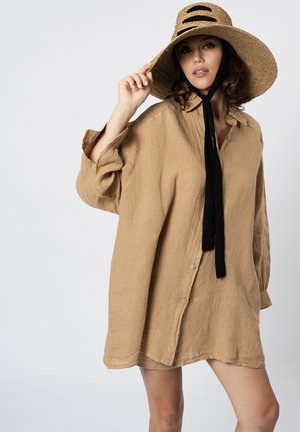Donna che indossa un vestito largo di lino beige e un cappello di paglia a tesa larga con nastro nero, posando davanti a uno sfondo chiaro e semplice.