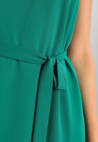 Vestido verde con una textura suave y fluida. Presenta un lazo en la cintura y un diseño sencillo, enfatizando la tela suave y el color vibrante.
