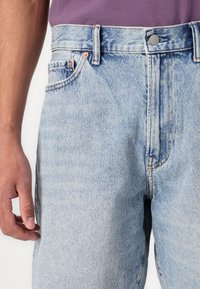 Jeans de mezclilla azul claro con cintura alta, diseño de cinco bolsillos y acentos de botón en tono cobre. Textura desgastada en la tela.