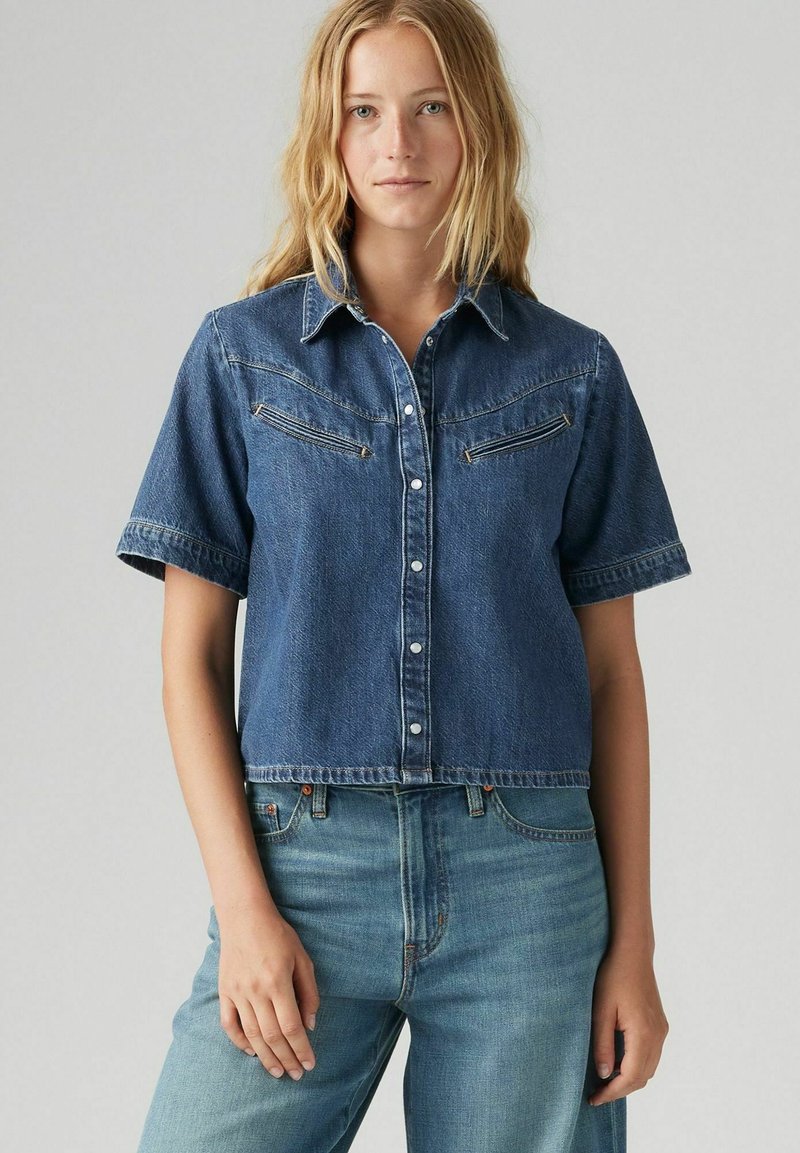 Camicia in denim di colore blu scuro, con maniche corte, collo a camicia, chiusura frontale con bottoni e due tasche sul petto. Tessuto morbido, vestibilità cropped.