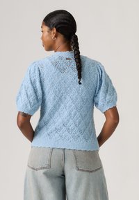 Mujer con el cabello trenzado que lleva un suéter de punto de manga corta con patrón en azul claro y jeans de talle alto en azul claro, vista de espaldas.