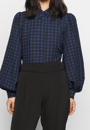 Vrouw draagt een blauw-zwart geruit overhemd met geplooide lange mouwen en een zwarte broek met hoge taille, staand tegen een effen achtergrond.