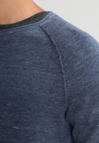 Mörkblå stickad tröja med texturerad tyg, med rund halsringning och raglanärmar, som visar ett subtilt prickigt mönster.