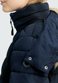 Person in einer marineblauen gesteppten Daunenjacke mit hohem Kragen und abnehmbarem Kapuze, die Druckknöpfe und Reißverschlüsse aufweist.