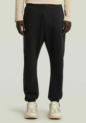 Personne portant un pantalon de jogging noir et des baskets beiges, debout devant un fond clair uni.