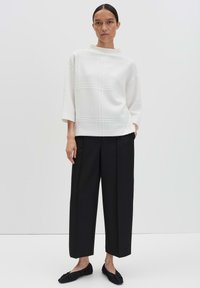 Donna che indossa un top bianco strutturato a tre quarti, pantaloni larghi neri a metà polpaccio e scarpe basse nere, in piedi davanti a uno sfondo semplice.