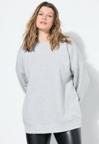 Mladá žena s vlnitými vlasy, oblečená ve světle šedé oversized mikině a černých kožených legínách na jednobarevném bílém pozadí.