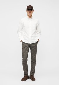 Νέος άνδρας που φορά λευκό πουκάμισο με κουμπιά, γκρι παντελόνι με σχέδιο, καφέ αθλητικά παπούτσια και καφέ καπέλο μπέιζμπολ, στέκεται με τα χέρια στις τσέπες.