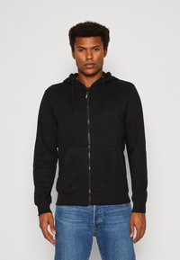 Schwarze Zip-Hoodie aus weichem Material, mit einer Kordelzugkapuze, Fronttaschen und einem dezenten Logo auf der Brust. Kombiniert mit Jeans.