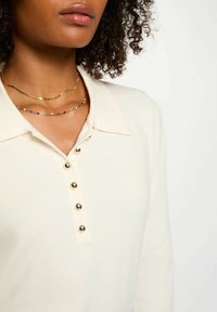 La polo bianca a maglia presenta un colletto e cinque bottoni metallici. Due collane sovrapposte: una in catena dorata, l'altra con perline dai colori vivaci.