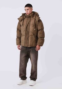 Chaqueta acolchada marrón con cuello alto, cierre frontal de cremallera y capucha con cordón, combinada con vaqueros oscuros y zapatillas blancas.