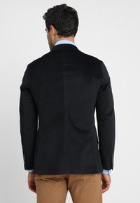 Svart kavaj med en strukturerad yta, enkel ventil bak, och kontrasterande ärmknappar. Buren över en ljusblå skjorta.