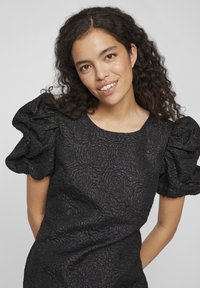 Μαύρο φόρεμα με υφασμάτινη υφή λουλουδιών, στρογγυλή λαιμόκοψη και φουσκωτά μανίκια. Το ύφασμα έχει ελαφρώς γυαλιστερό φινίρισμα.