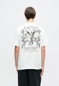 Biele tričko s krátkym rukávom s čiernym grafickým potlačou znázorňujúcou okrídlenú postavu, "NIKE" a dekoračné prvky na chrbte.