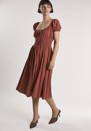 Frau in einem rostfarbenen, knielangen Kleid mit Puffärmeln und geraffter Taille, die gewebte, hochhackige Mules trägt, vor weißem Hintergrund.