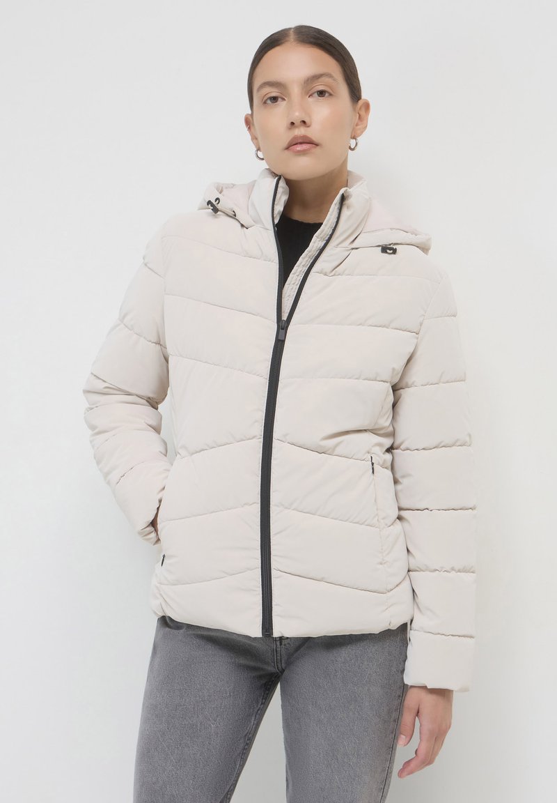 Frau trägt eine hellbeige gesteppte Jacke mit Kapuze, die bis oben zugezippt ist, mit den Händen in der Tasche, kombiniert mit grauen Jeans auf weißem Hintergrund.