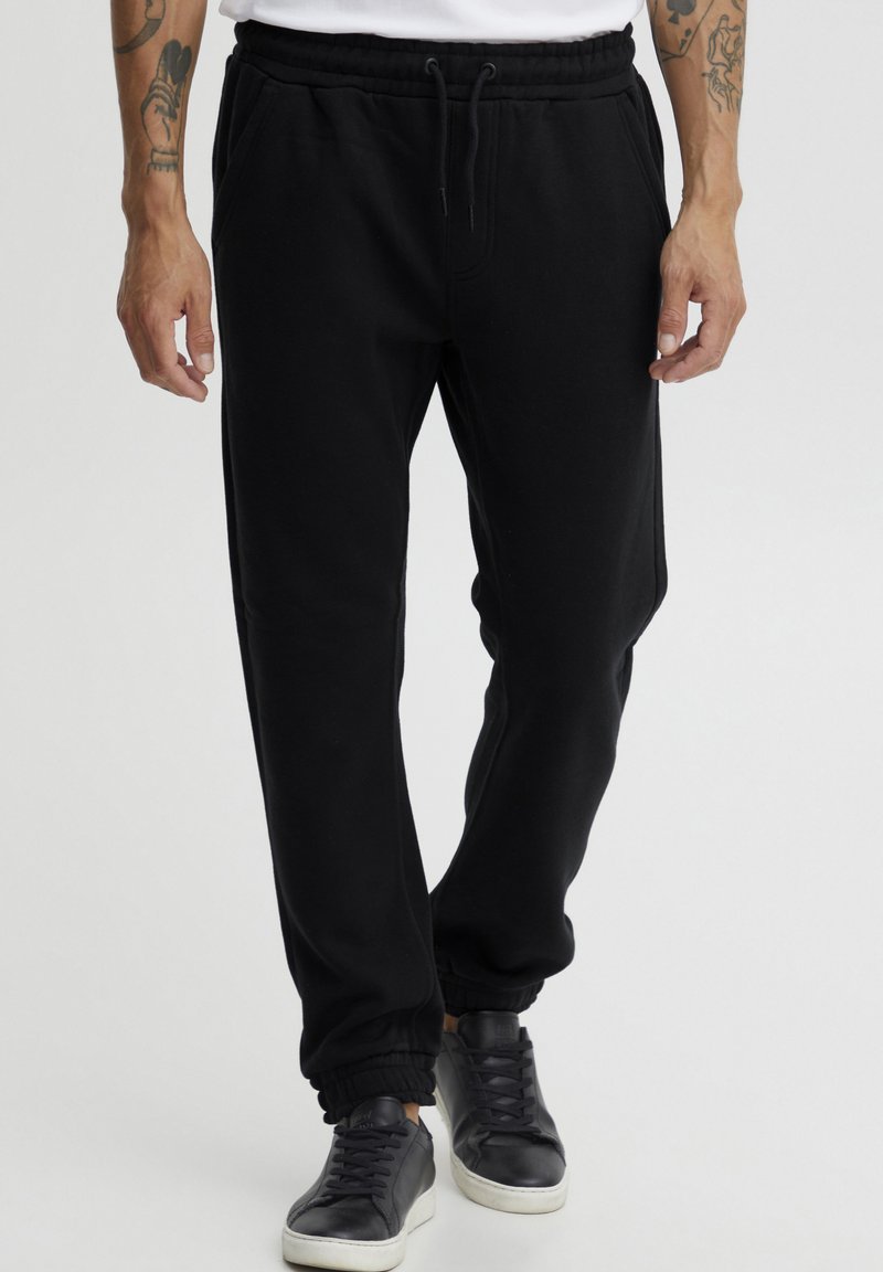 Pantalones de chándal negros hechos de tela suave, con una cintura elástica, cordón ajustable y puños ajustados. Se llevan con zapatillas negras.