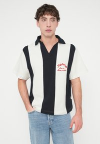 Κοντομάνικο πόλο με γιακά, με έντονες κάθετες ρίγες σε μαύρο και άσπρο, και κεντημένο κόκκινο κείμενο στην αριστερή πλευρά του στήθους.