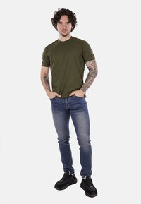 T-shirt di cotone verde oliva abbinato a jeans blu a vita alta. L'outfit è completato da scarpe sportive nere e presenta braccia tatuate.