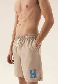 Beżowe szorty kąpielowe z perforowanej tkaniny, elastycznym pasem i białym sznurkiem. Zawierają niebieską literę "H" po lewej stronie.
