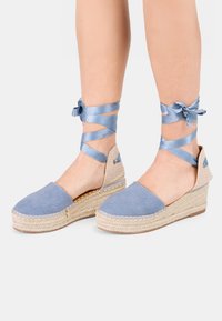 Modré plátno espadrilky so zavedenými espadrilkovými podpätkami z juty. Má šnúrky na členkoch na zapínanie a jemne textúrovaný zadný panel.
