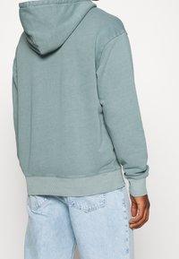 Huvtröja i dämpad tealfärg, gjord av mjukt tyg. Har en avslappnad passform, ribbade muddar och fåll. Kombinerad med ljusblå jeans.