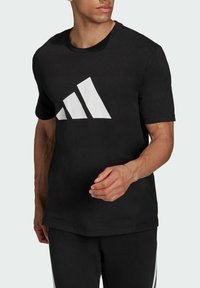 Mustat puuvillapaidat, joissa on suuri valkoinen Adidas kolme nauhaa -logo etupuolella, lyhyet hihat ja pyöreä kaula-aukko.