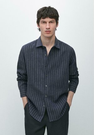 Giovane uomo con capelli scuri che indossa una maglietta a maniche lunghe blu navy scuro con righe verticali bianche, in piedi con le mani in tasca davanti a uno sfondo semplice.