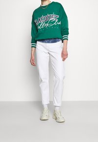 Sudadera verde con texto blanco y detalles en azul marino, combinada con pantalones blancos ajustados y zapatillas deportivas beige. Diseño sencillo y corte relajado.