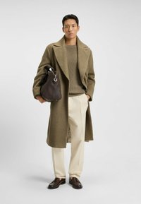Olive green wool coat, beige ribbed sweater, cream tailored pants, brown leather shoes, holding a dark brown handbag with silver hardware.