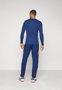 Completo sportivo da uomo in blu scuro con maniche lunghe e pantaloni coordinati, caratterizzato da accenti arancioni e un design aderente. Il materiale sembra liscio.