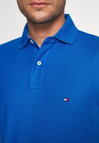 Modrý polo tričko vyrobené z texturovaného materiálu, vybavené límcem a dvěma knoflíky. Mezi pozoruhodné detaily patří malá vyšitá nášivka v červené, bílé a modré barvě.
