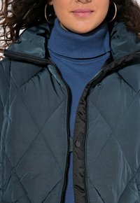 Giacca puffer trapuntata in verde scuro con colletto alto e zip nera. Indossata sopra un dolcevita blu, che mette in risalto una texture liscia.