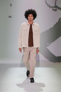 Model îmbrăcat cu o jachetă ușor gri, cu butoni, peste o cămașă maro, pantaloni bej și pantofi negri, plimbându-se pe un podium cu un fundal simplu.