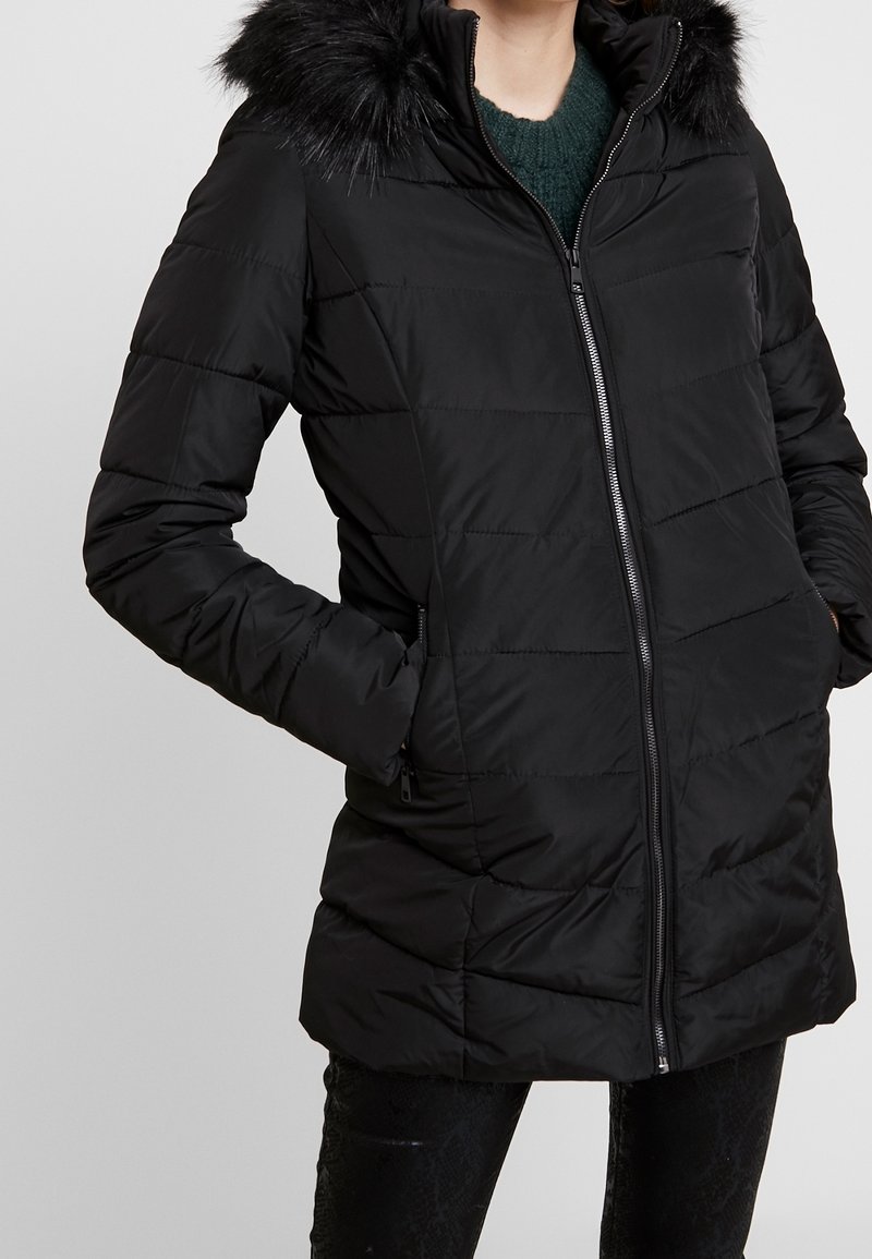 Doudoune noire avec zip sur le devant, capuche ornée de fausse fourrure et design à panneaux cousus. Associée à un pantalon noir texturé.