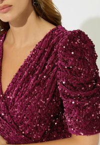 Robe en sequins bordeaux avec des manches texturées et bouffantes ; décolleté en V profond ; superposition complexe de sequins créant une surface riche et réfléchissante.
